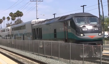 Un tren Metrolink gris oscuro y verde con número 923 en vías, junto a una valla. Al fondo se aprecian palmeras, postes de energía y un cielo azul