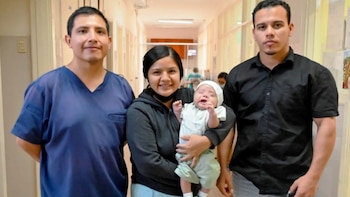 Hombre con uniforme médico azul, mujer con bebé en brazos, hombre de camisa negra posan en un pasillo de hospital