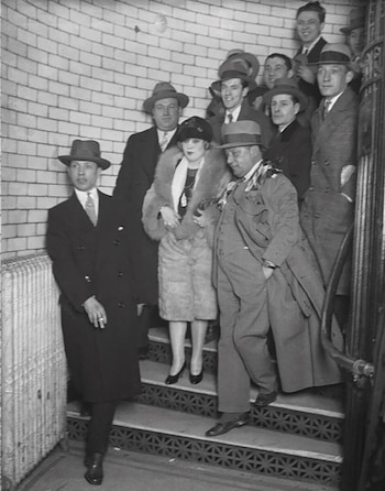 Fotografía en blanco y negro de Mae West con un abrigo de piel y sombrero, descendiendo una escalera con varios hombres con traje y sombrero