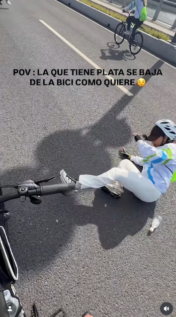 Una mujer con casco y ropa blanca en el suelo de una vía junto al manillar de una bicicleta y un ciclista en movimiento al fondo