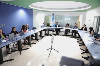 Vista amplia de una sala de conferencias moderna con paredes azules y blancas, donde varias personas están sentadas alrededor de una mesa grande en forma de U
