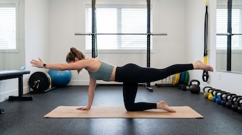 Mujer realizando el ejercicio bird dog sobre una colchoneta en un gimnasio. Se apoya en rodilla y mano, con el brazo derecho y la pierna izquierda extendidos.