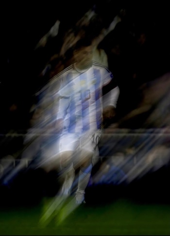 Hombre con camiseta de fútbol de Argentina con rayas celestes y blancas y el número diez, en movimiento borroso, sobre un fondo oscuro con tonos rojos