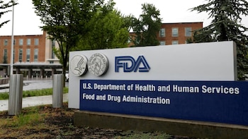 Primer plano del cartel blanco de la FDA con letras azules y los logotipos del DHS, frente a un edificio de ladrillo rojo y árboles verdes bajo un cielo nublado