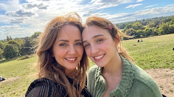 Dos mujeres en primer plano sonríen hacia la cámara en una pradera verde con árboles y cielo con nubes de fondo.