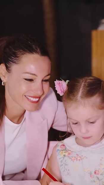 Una mujer de cabello recogido y blazer rosa sonríe junto a una niña con coletas y vestido floral, que sostiene un objeto rojo y mira hacia abajo
