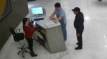 Una mujer con teléfono, un hombre detrás de un mostrador y una persona de pie. Hay un monitor de computadora y una silla.