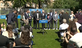 Grupo de personas, mayormente hombres de traje, en un césped verde con un podio 'FIFA arena' y público sentado. Una estructura con carteles de FIFA de fondo