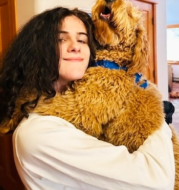 Una persona joven con cabello oscuro y rizado, vistiendo una camisa blanca, abraza a un perro marrón peludo con un collar azul. El perro mira hacia arriba con la boca abierta