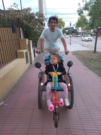 Un hombre empuja un triciclo rojo y azul con una niña pequeña sentada en él por una acera de ladrillo. Ambos miran al frente. Hay árboles y coches al fondo