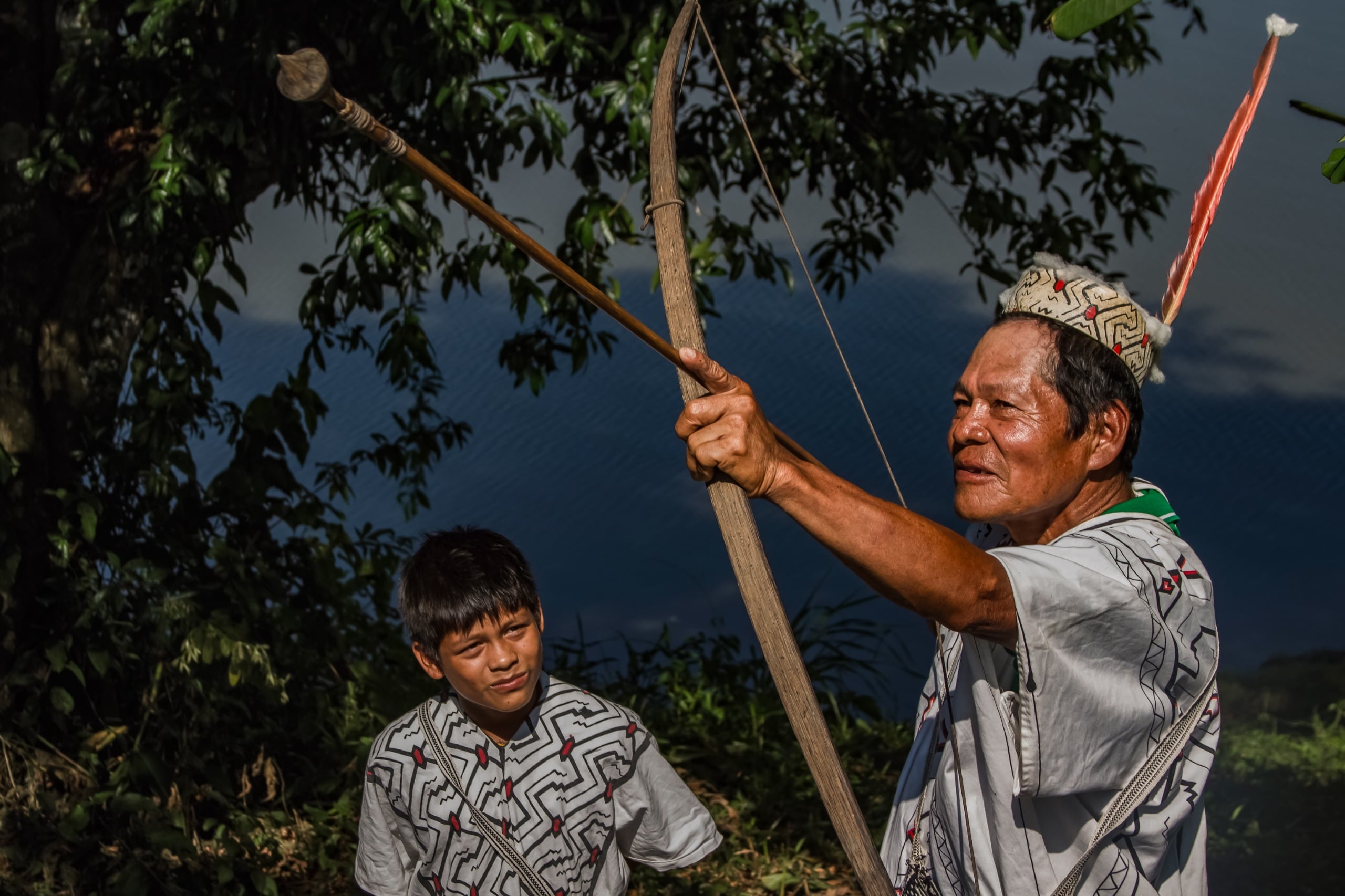 Un hombre indígena mayor, ataviado con vestimenta tradicional y un tocado de plumas, sostiene un arco y una flecha mientras un joven lo observa atentamente, en una imagen que evoca la transmisión cultural y la vida en armonía con la naturaleza. (MIDIS)