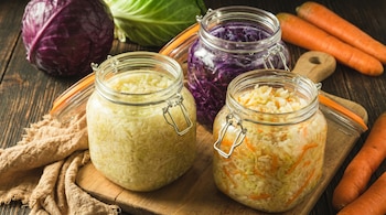 Tres frascos de vidrio con col fermentada, dos de col blanca y uno de col morada, sobre una tabla de madera. Hay repollo fresco y zanahorias al fondo.