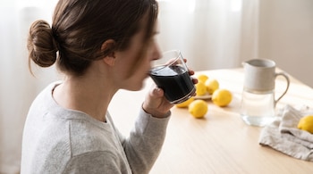 Una persona de perfil bebe de un vaso con líquido negro. Cabello castaño recogido, suéter gris claro. Limones y jarra de agua sobre la mesa.
