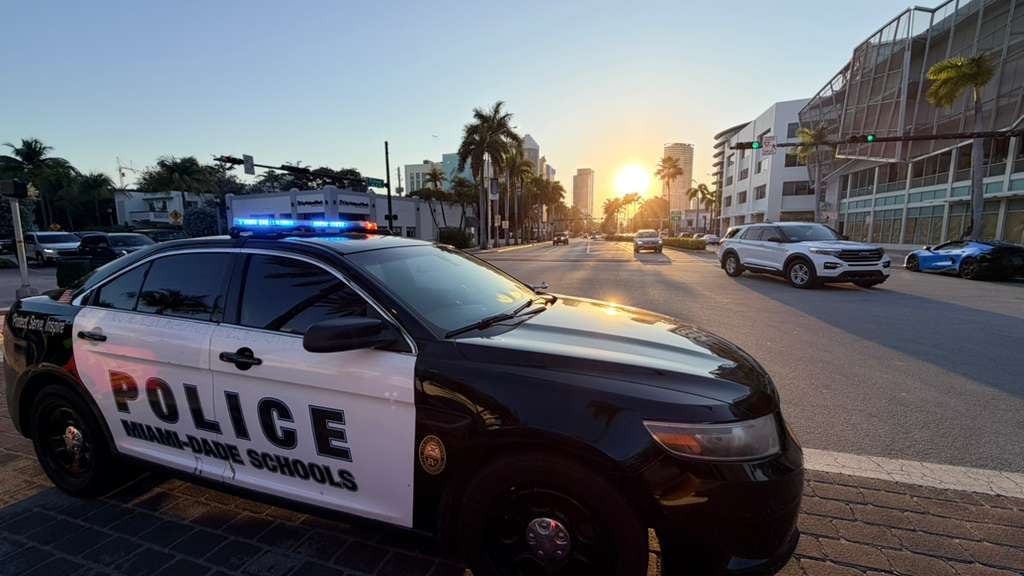 Un vehículo de la Policía Escolar de Miami-Dade se encuentra estacionado en una calle urbana mientras el sol se pone sobre edificios y palmeras, con un SUV y un auto deportivo visibles en la distancia (Opy Morales)