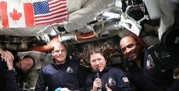 Cuatro astronautas flotan en una nave espacial. Christina Koch sostiene un micrófono, y banderas de Canadá y Estados Unidos cuelgan