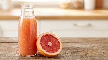 Una botella de vidrio transparente con jugo de color anaranjado y medio pomelo rosado sobre una mesa de madera.
