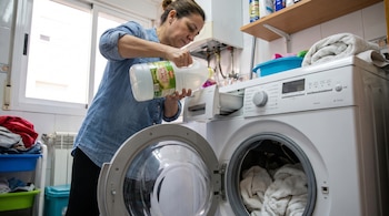 Una mujer de mediana edad con camisa vaquera vierte vinagre blanco en el dispensador de una lavadora que contiene toallas.