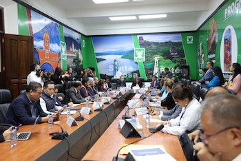 Personas sentadas alrededor de una mesa con micrófonos y letreros mientras un grupo de periodistas toma fotografías