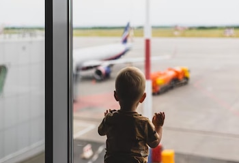 Vista trasera de un niño pequeño de cabello claro mirando por una ventana grande hacia la pista de un aeropuerto con un avión blanco y un vehículo naranja de fondo