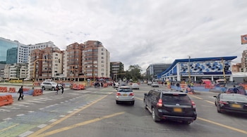 Intersección de calle urbana con vehículos, peatones en el paso de cebra, semáforos, barreras naranjas y edificios al fondo.
