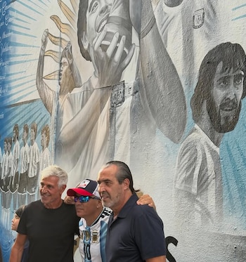 Tres hombres posan frente a un gran mural. El mural presenta a Diego Maradona sosteniendo un trofeo, otros futbolistas argentinos y nombres escritos. Los hombres sonríen