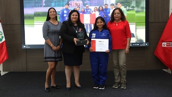 Cuatro personas, tres mujeres adultas y una adolescente con mono azul, posan en un escenario. Una mujer sostiene un trofeo, la adolescente un diploma. Bandera de Perú visible
