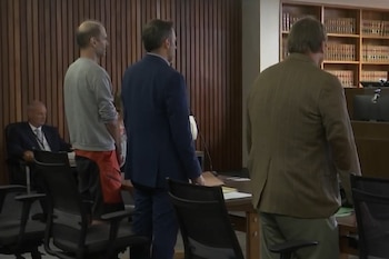 Un hombre con sudadera gris y pantalones cortos rojos permanece de pie junto a dos hombres en traje dentro de una sala de tribunal