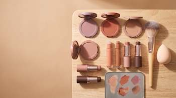 Rubores compactos, tubos de maquillaje, barra cremosa, brocha, esponja y paleta con muestras en una tabla de madera.