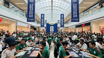Multitud de personas sentadas en mesas intercambiando estampas del Mundial 2026 en un centro comercial con techo de cristal y banderas azules colgantes.