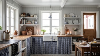 Cocina con paredes y techo de paneles blancos, ventanas, un fregadero, cortinas a cuadros azules, estantes con cerámica, una estufa antigua y una mesa de madera con desayuno.