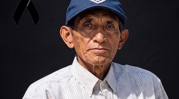 Primer plano de un hombre anciano con gorra azul y camisa a rayas, sobre fondo negro con un crespón negro en la esquina superior izquierda.