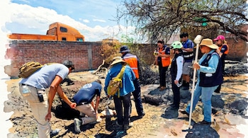 Varios individuos con chalecos de seguridad examinan un terreno árido y rocoso. Uno se inclina hacia un agujero; un camión naranja y pared de ladrillos al fondo.
