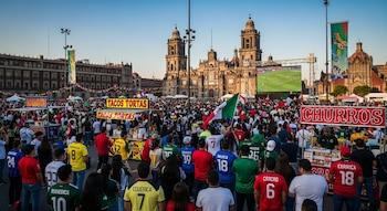 Miles de personas en el Zócalo de México con camisetas de fútbol, mirando una pantalla gigante con un partido. La Catedral Metropolitana y puestos de comida son visibles.