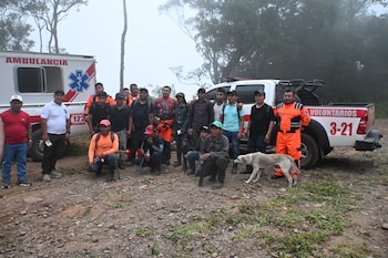 Un grupo de dieciocho hombres posa junto a una ambulancia blanca y una camioneta pick-up de voluntarios, con un perro claro al frente en un terreno de tierra