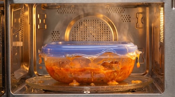 Primer plano del interior de un microondas con un recipiente plástico transparente y tapa azul derritiéndose, revelando comida naranja burbujeante y vapor.