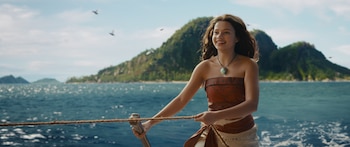 Catherine Laga'aia como Moana en un bote. Viste un top y falda marrones, un collar turquesa, y sonríe mientras sujeta una cuerda con montañas de fondo
