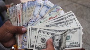 Dos manos sujetan varios billetes de soles peruanos desplegados en abanico junto a fajos de billetes de cien dólares estadounidenses