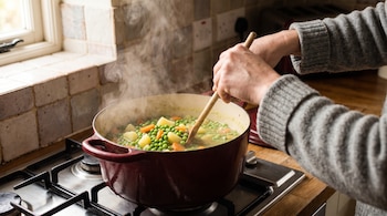 Brazos con suéter gris sostienen una cuchara de madera que revuelve un guiso de arvejas en una olla roja sobre una cocina.