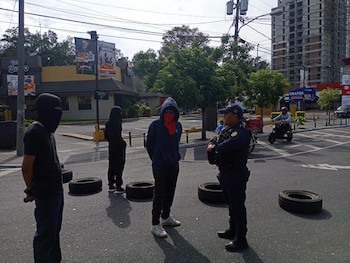Tres personas con el rostro cubierto y un agente de policía de pie en una calle con varios neumáticos. Al fondo, edificios y árboles