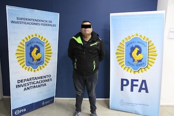 Hombre de pie con los ojos censurados, vestido con chaqueta negra y jeans, flanqueado por dos pancartas de la Policía Federal Argentina