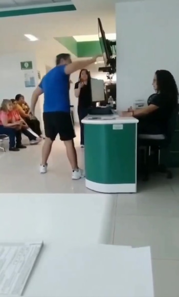 Un hombre en camiseta azul y shorts oscuros gesticula con los brazos abiertos frente a una empleada sentada detrás de un mostrador verde en una sala de espera