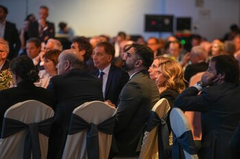 Varias personas vestidas formalmente sentadas en mesas de evento. Un hombre con barba oscura mira hacia la derecha, flanqueado por dos mujeres