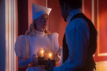Mujer de cabello rubio con vestimenta blanca de época y sombrero cónico sostiene dos linternas con velas encendidas frente a un hombre. Fondo rojo