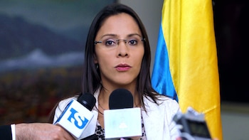 Una mujer con gafas y chaqueta clara habla ante varios micrófonos con la bandera de Venezuela al fondo