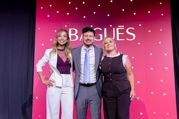 Tres personas sonrientes en un escenario con fondo rosa y el logo Baguēs. Una mujer de blanco, un hombre de traje gris, y una mujer con chaleco oscuro