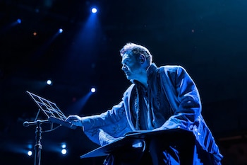 Un hombre de perfil derecho está de pie en un escenario oscuro, con iluminación azul, junto a un atril y una superficie plana. Viste un traje oscuro