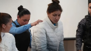 Mujer con chaqueta acolchada clara y otra mujer con uniforme azul que le toca el hombro. Un hombre de uniforme oscuro visible a la derecha