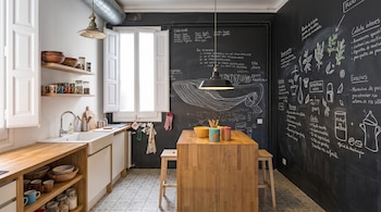 Cocina con pared de pizarra decorada con dibujos de tiza, una mesa isla de madera, dos taburetes, fregadero, ventanas y estantes con menaje de cocina.