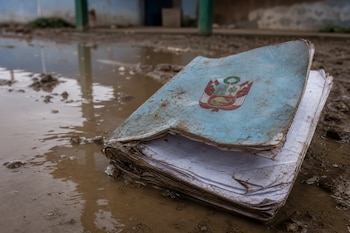 Un cuaderno azul con el escudo de Perú, manchado de lodo y con las hojas mojadas, sobre un charco de agua en el suelo.