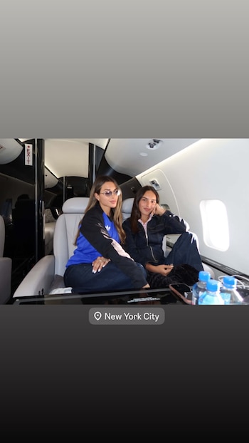 Dos mujeres sentadas en los asientos de un avión privado. La cabina tiene una señal de salida. Hay botellas de agua y un teléfono sobre una mesa lateral